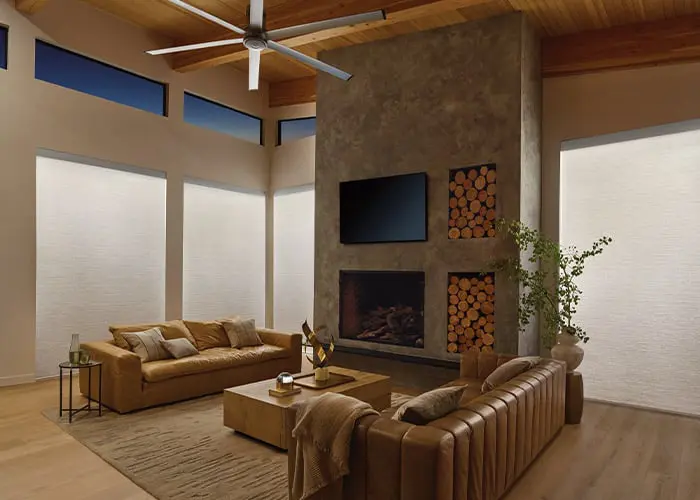 Spacious living room featuring a contemporary stone fireplace, high ceilings with wooden beams, large windows with blinds, and cozy leather sofas. Elegant, bright, with natural lighting.