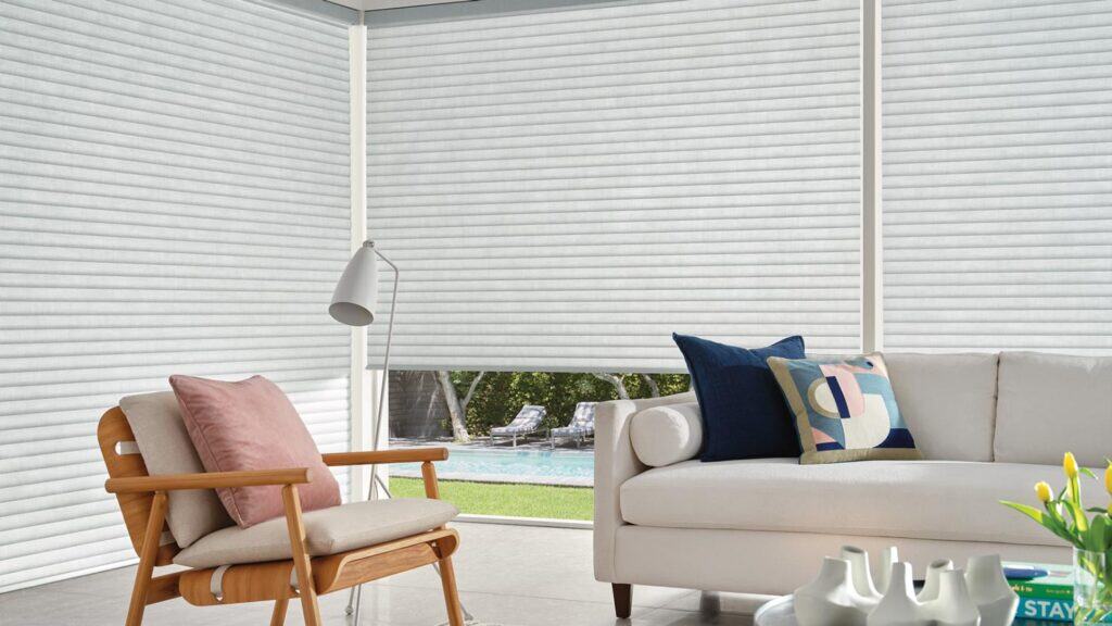 Bright indoor sunroom with white window blinds, comfortable white sofa with colorful pillows, wooden lounge chair, and view of outdoor pool and garden. Contemporary style, natural light, relaxing atmosphere, ideal for leisure and entertaining.